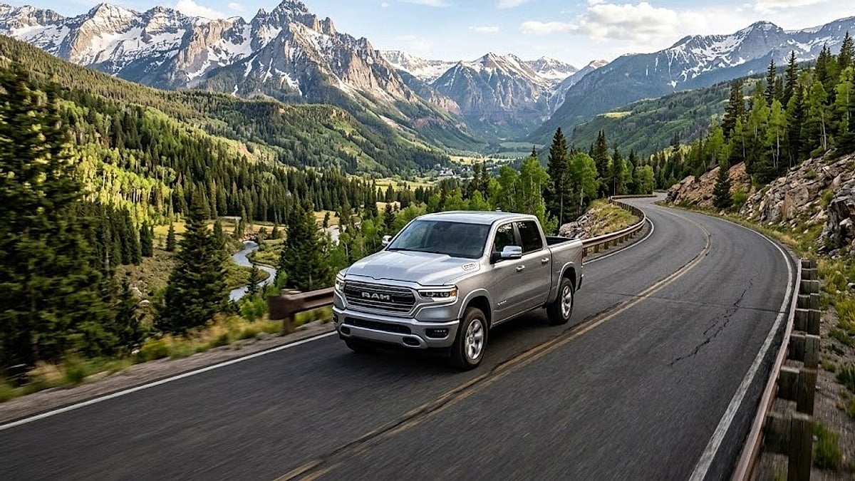 A silver 2026 Ram 1500 drives along a winding Colorado mountain road with massive, snow-capped rocky peaks and forests