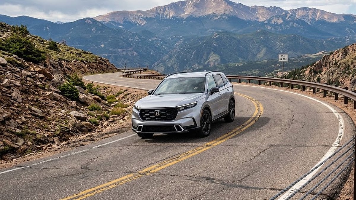 A silver Honda CR-V travels along a winding mountain road with a large peak and valleys in the background