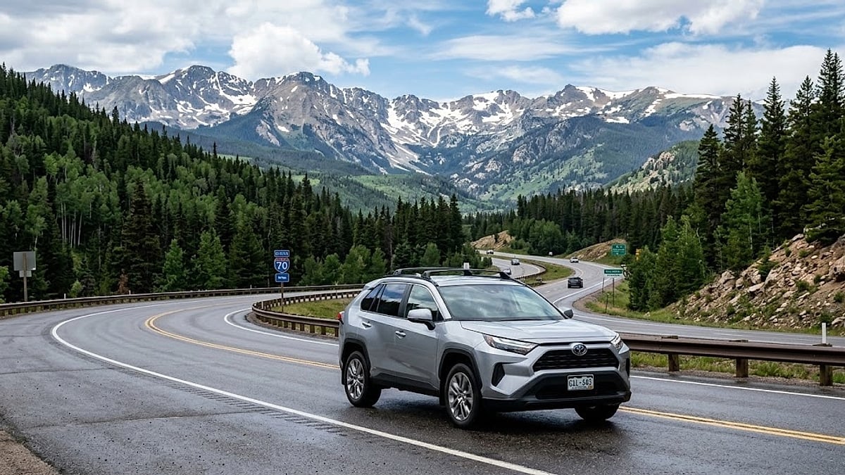 A silver Toyota RAV4 hybrid drives along a winding mountain highway in the Colorado Rockies, with I-70 signs and snow-capped peaks