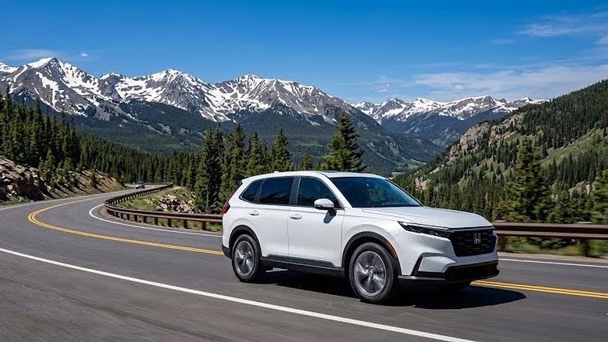 A white 2026 Honda CR-V Hybrid drives along a winding asphalt mountain highway curve in front of snow-capped peaks