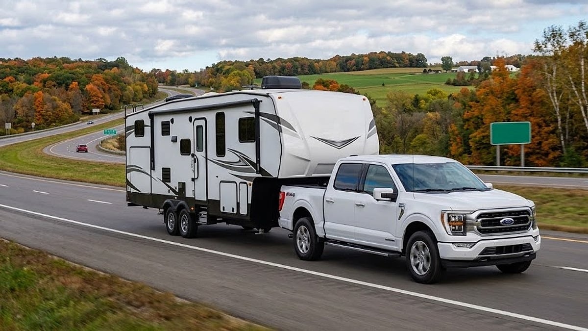 A white Ford F-150 towing a large white travel trailer with a prominent air conditioner drives down a multi-lane highway surrounded by vibrant, colorful autumn forests and distant farm fields on a cloudy day
