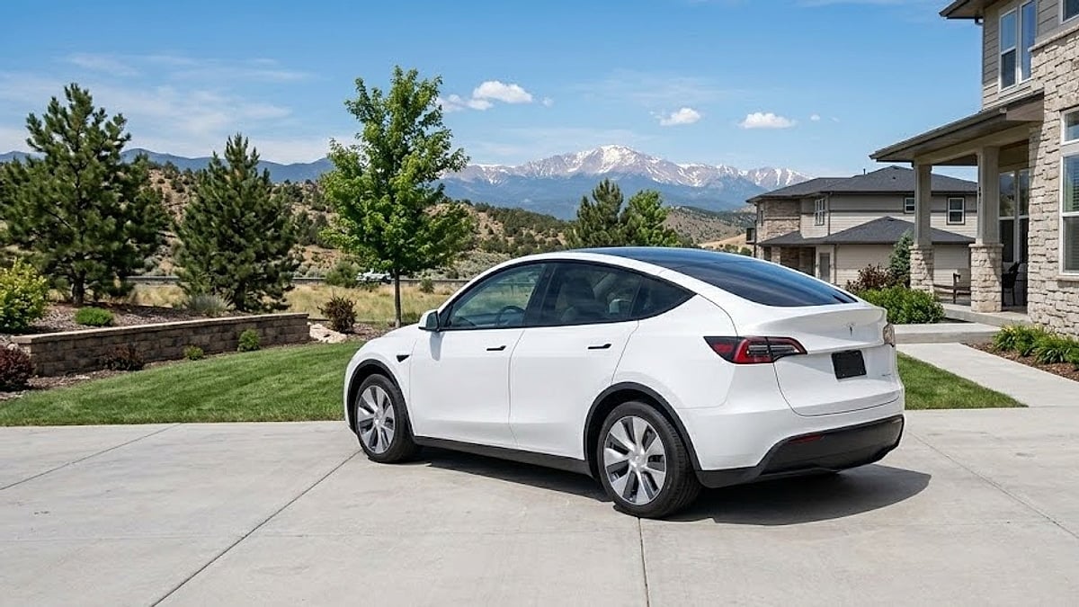 A white Tesla Model Y sits parked in a concrete suburban Colorado driveway, framed by manicured landscaping and mountain peaks