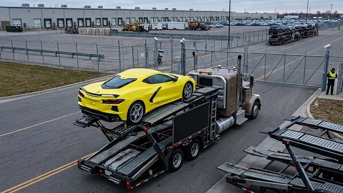 A yellow 2025 Chevrolet Corvette sits loaded on a flatbed transport carrier entering a secure industrial automotive inspection facility