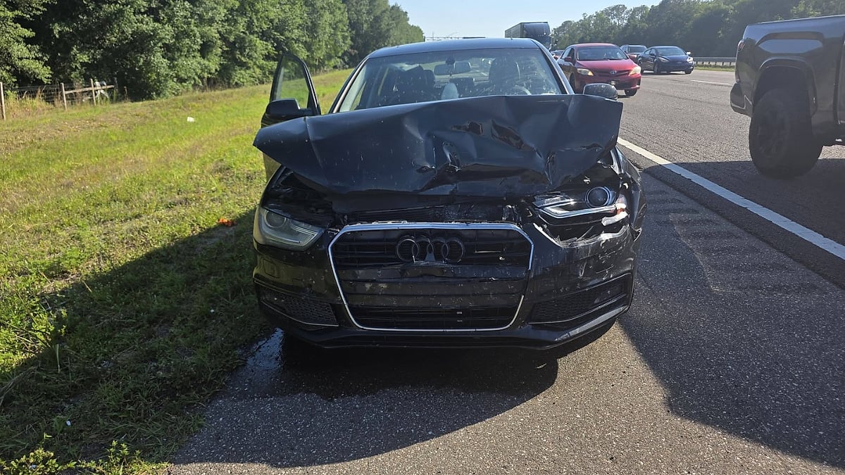 Front view of a black Audi A4 with severe front-end damage after a highway collision involving a Rivian R1S.