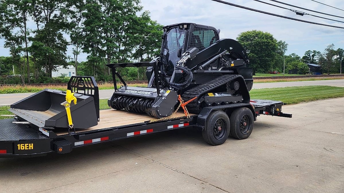Black skid steer loaded on a Big Tex equipment trailer parked on a concrete lot.
