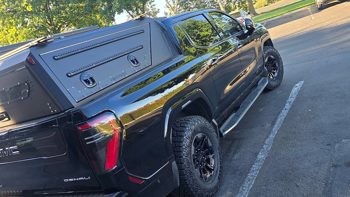 Black GMC Sierra Denali with an aftermarket bed cap shown from the rear side in a parking lot.