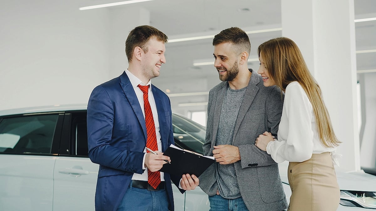 A car salesperson speaks with a smiling couple while holding paperwork inside a bright dealership showroom.