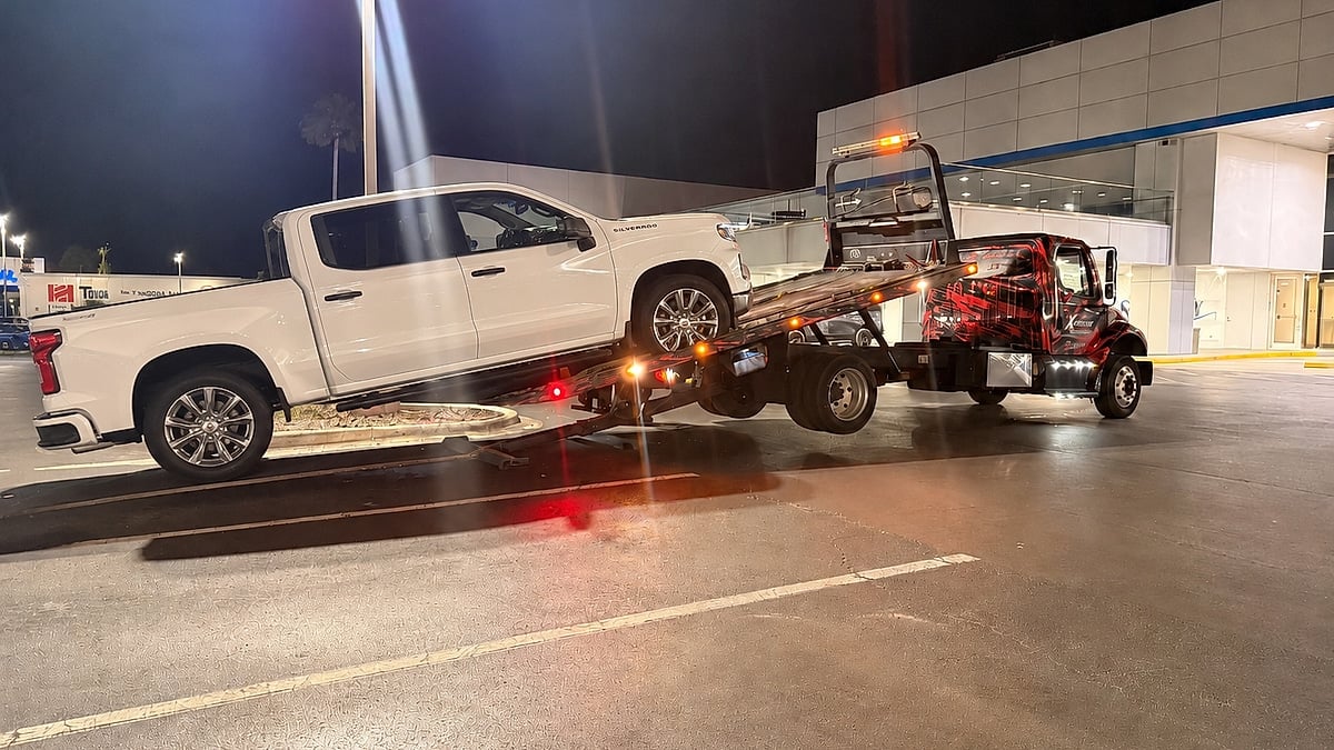 White Chevrolet Silverado being loaded onto a flatbed tow truck at night outside a dealership.
