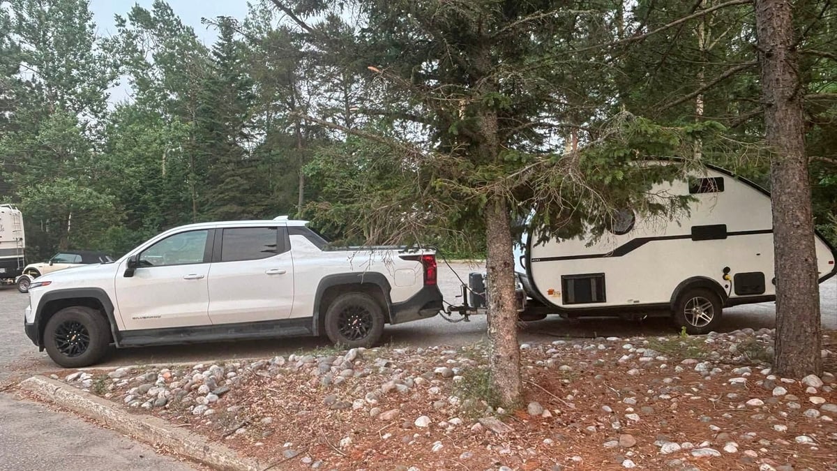 White Chevrolet Silverado EV towing a small white teardrop camper trailer, parked at a forested campsite