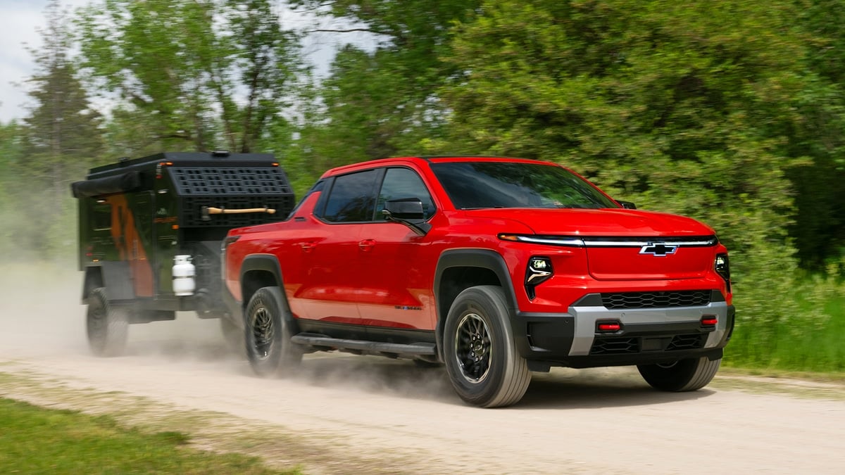 Red Chevrolet Silverado EV Trail Boss towing a black off-road camper trailer on a dusty dirt road through the woods
