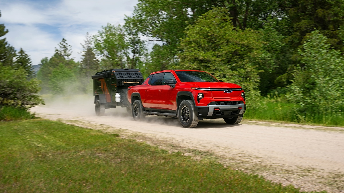 Red Chevrolet Silverado EV Trail Boss towing a small trailer along a dusty dirt road bordered by green trees.