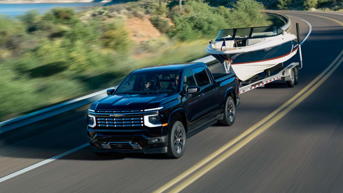 Black Chevrolet Silverado High Country pickup towing a boat on a coastal mountain road beside a lake