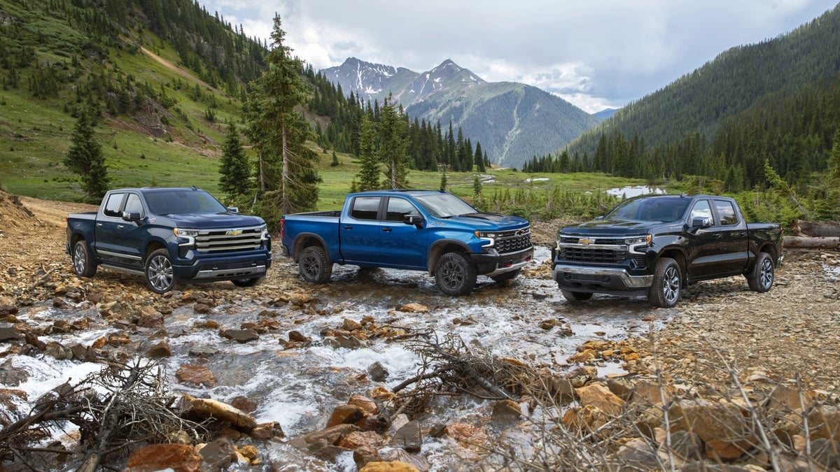 Three Chevrolet Silverado pickup trucks in dark blue, bright blue, and black parked in a rocky mountain stream
