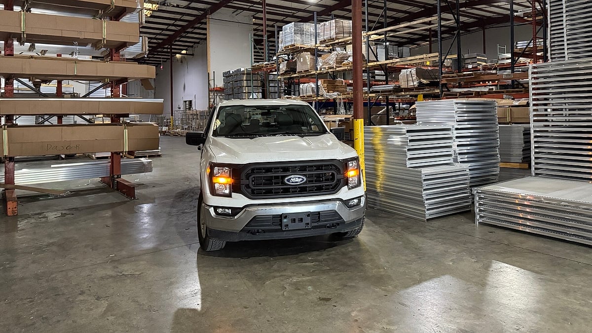 White Ford F-150 parked inside a warehouse surrounded by stacked metal panels and shelving.