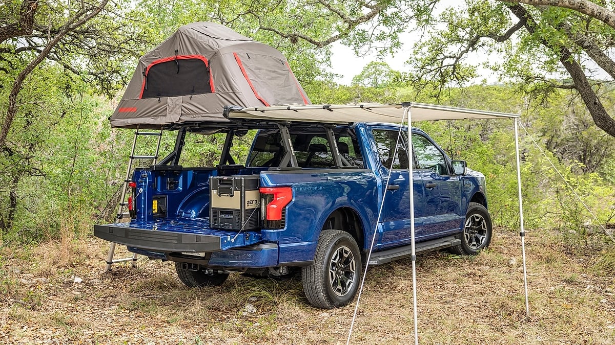 Blue Ford F-150 Lightning set up for camping with a rooftop tent, awning, and gear in the bed.