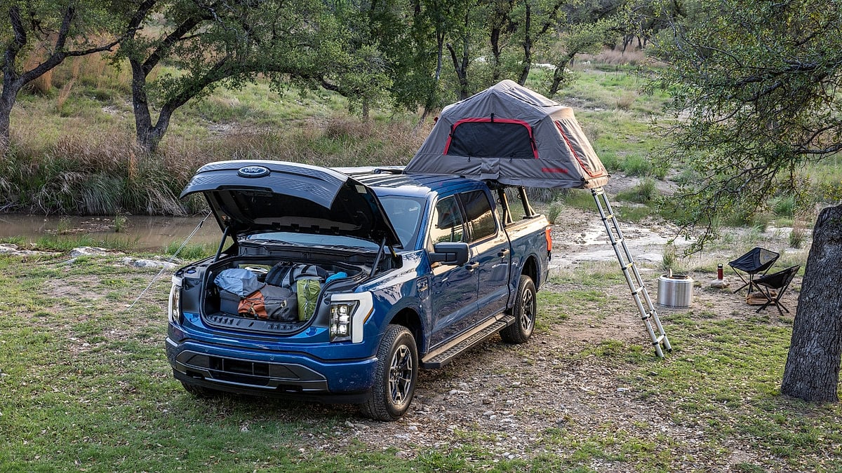 Blue Ford F-150 Lightning XLT set up at a campsite with an open frunk and rooftop tent.