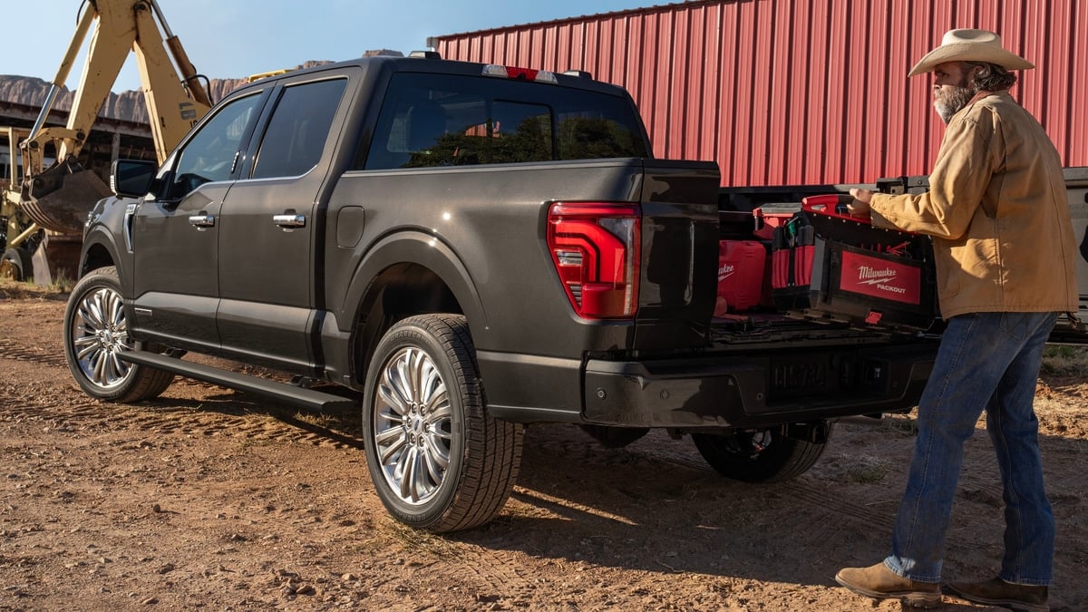 Dark gray Ford F-150 Platinum pickup with tailgate open, a man loading a Milwaukee Packout tool box at a rural jobsite