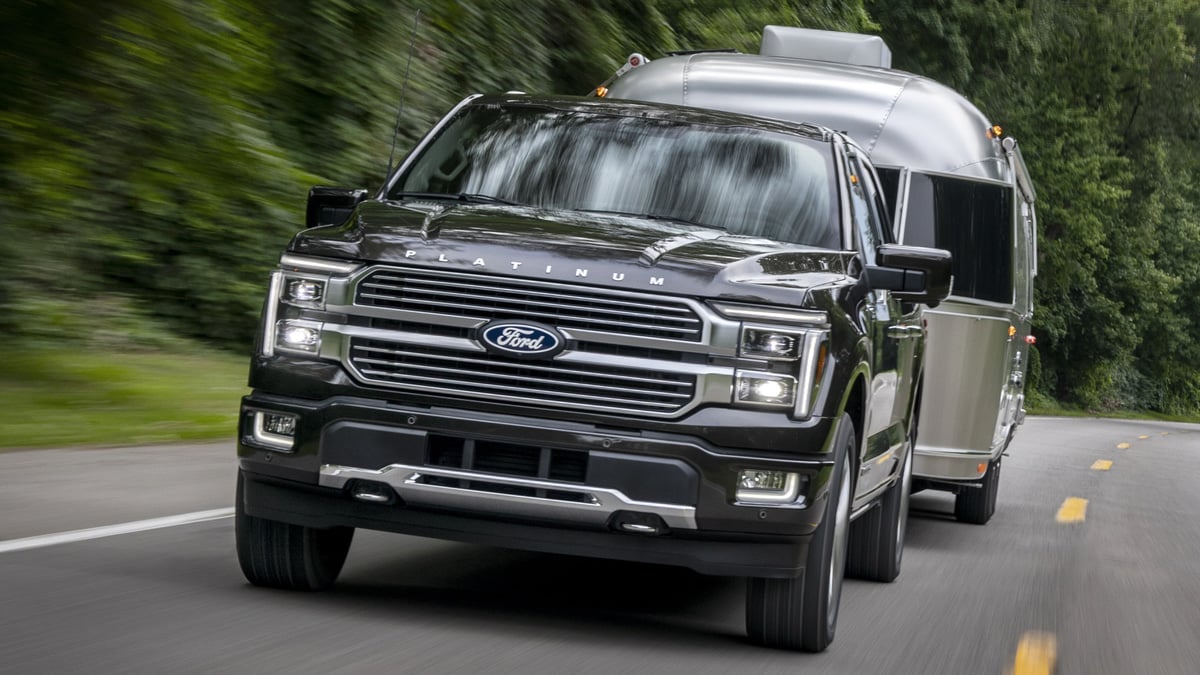 Dark gray Ford F-150 Platinum pickup towing a silver Airstream travel trailer on a tree-lined road, front three-quarter view