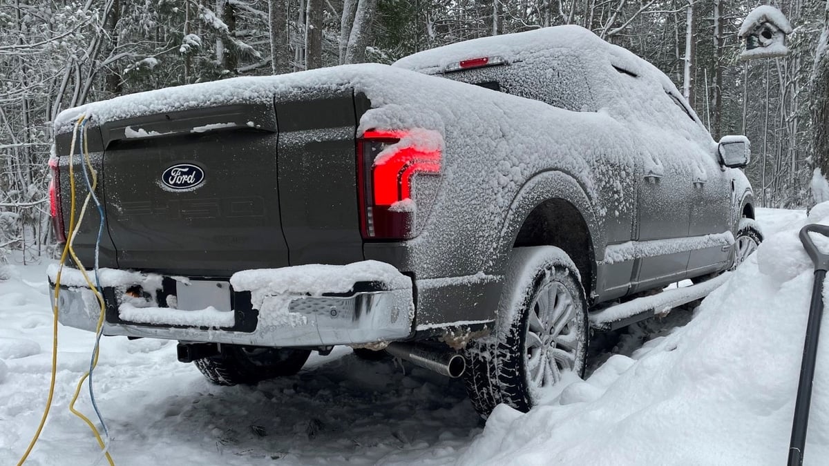 Snow-covered Ford F-150 pickup truck parked in a snowy forest, rear view with taillights lit and a snow shovel nearby