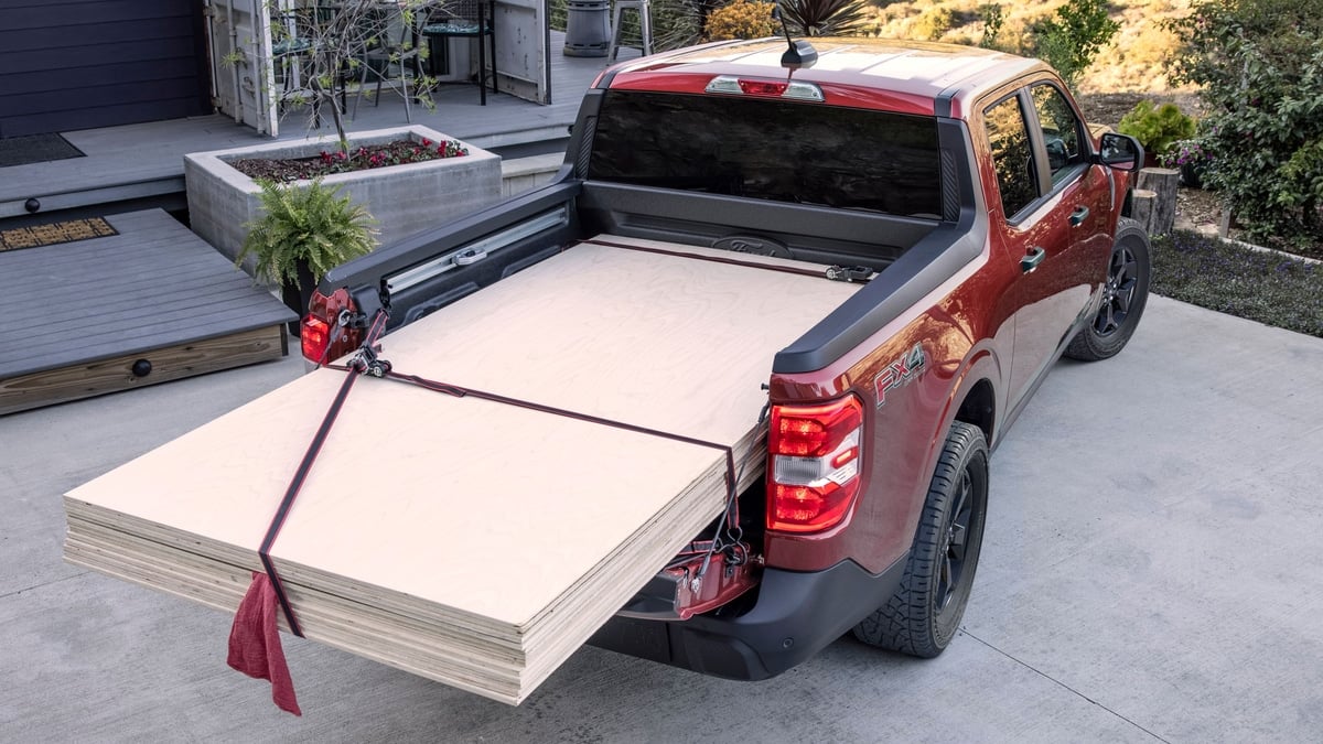 Red Ford Maverick FX4 pickup with its FlexBed extended, hauling sheets of plywood strapped down in a driveway