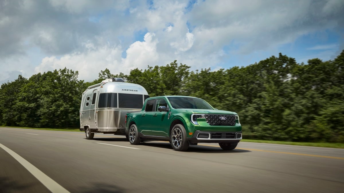 Green 2025 Ford Maverick towing an Airstream travel trailer on a highway in a front three-quarter action view.