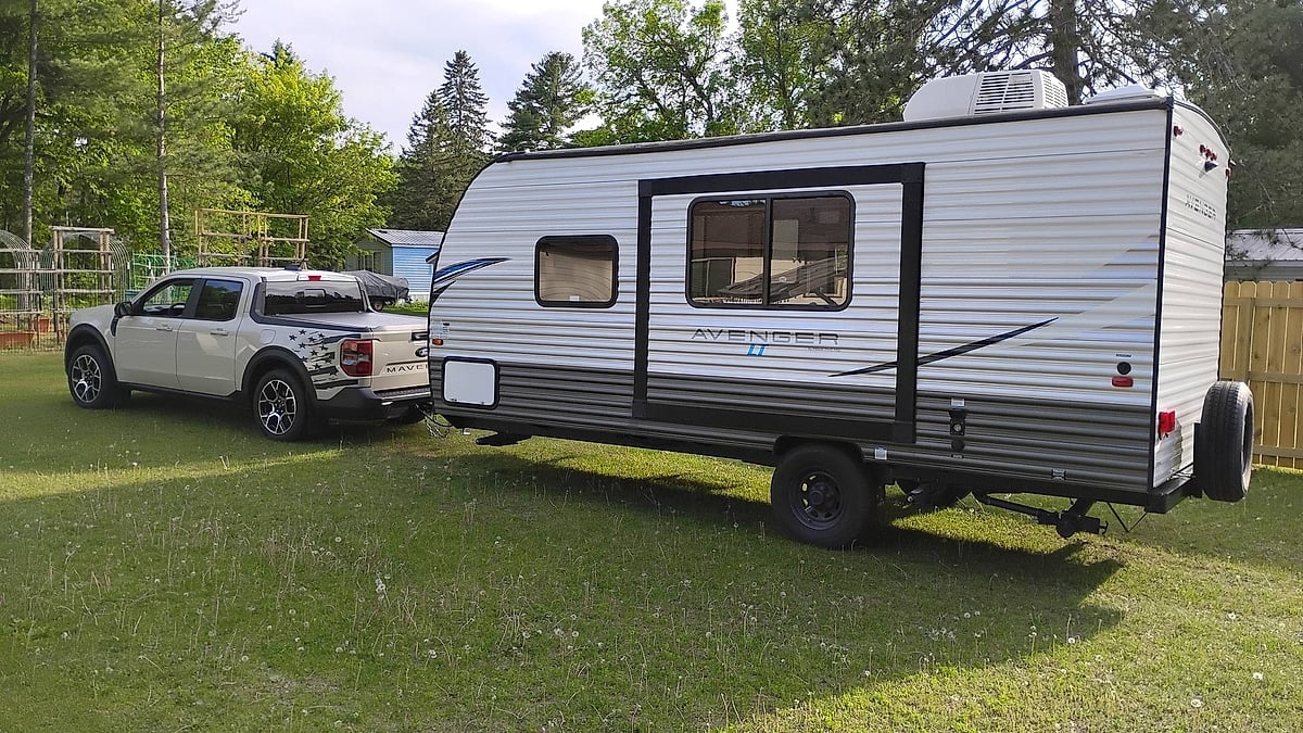 White Ford Maverick towing an Avenger LT travel trailer in a backyard side view.