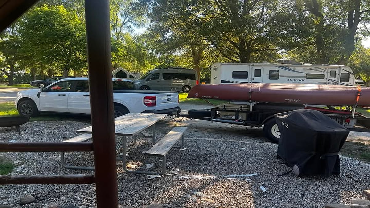 White Ford Maverick towing a canoe on a small trailer at a campsite with an Outback RV in the background.