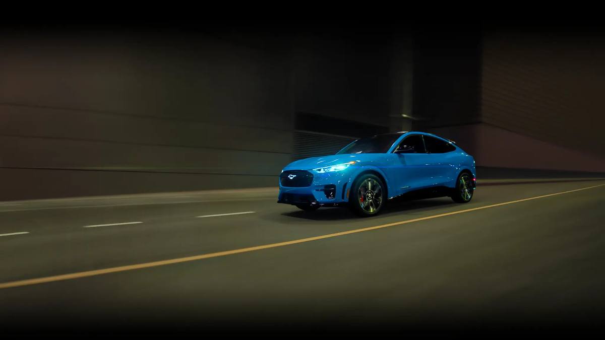 Blue Ford Mustang Mach-E GT electric SUV driving through a city at night with headlights on.