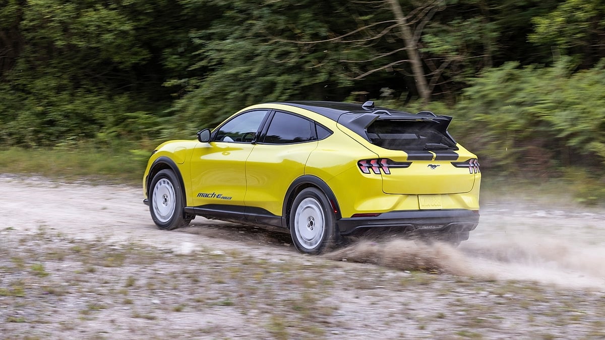 Yellow Ford Mustang Mach-E Rally driving on gravel and kicking up dust in a rear three-quarter action view.