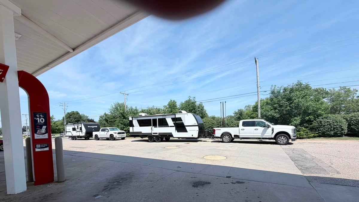 White Ford Super Duty pickup towing a large travel trailer at a gas station, with another truck and trailer parked nearby
