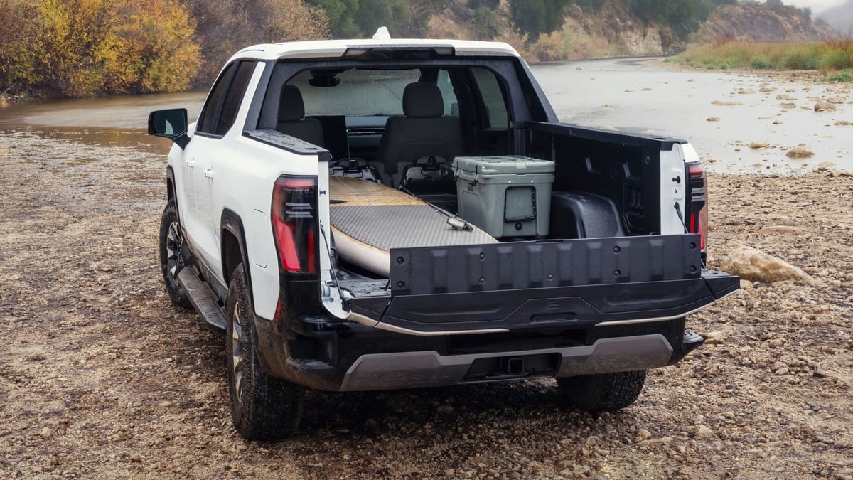White GMC Sierra EV pickup with its MultiPro midgate open, showing a paddleboard and cooler loaded in the bed near a riverbed
