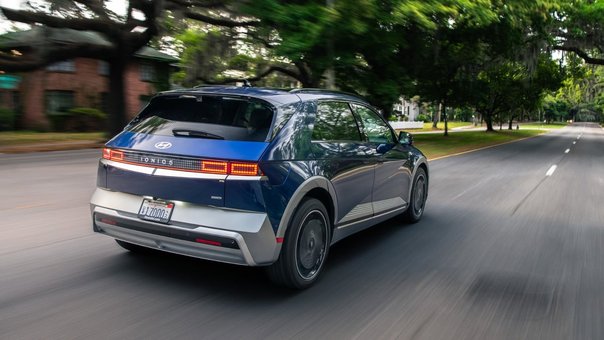 Blue Hyundai Ioniq 5 driving along a tree-lined road in a rear three-quarter action view.