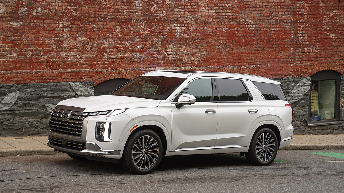 White Hyundai Palisade SUV parked in profile along a city street in front of a brick wall.