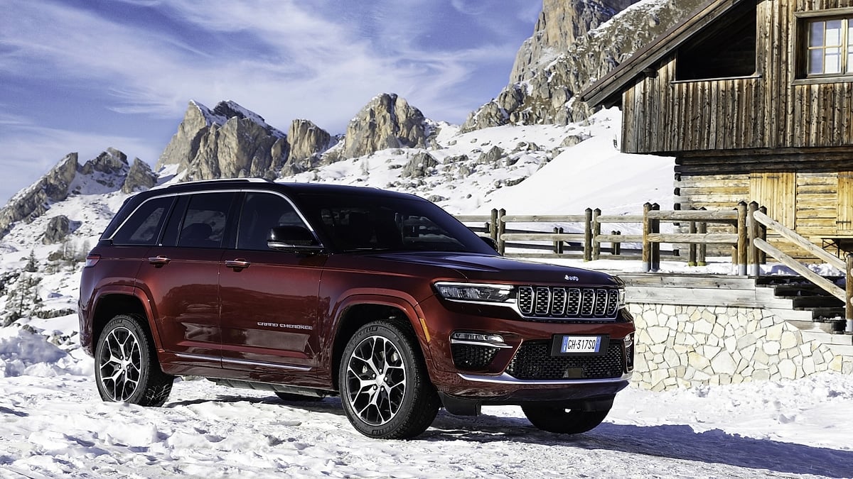 Burgundy Jeep Grand Cherokee 4xe parked in snow near a wooden cabin with rugged mountains in the background.