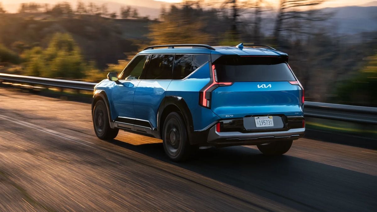 Blue Kia EV9 electric SUV driving on a highway, rear view showing aerodynamic design, LED taillights, and modern EV styling.