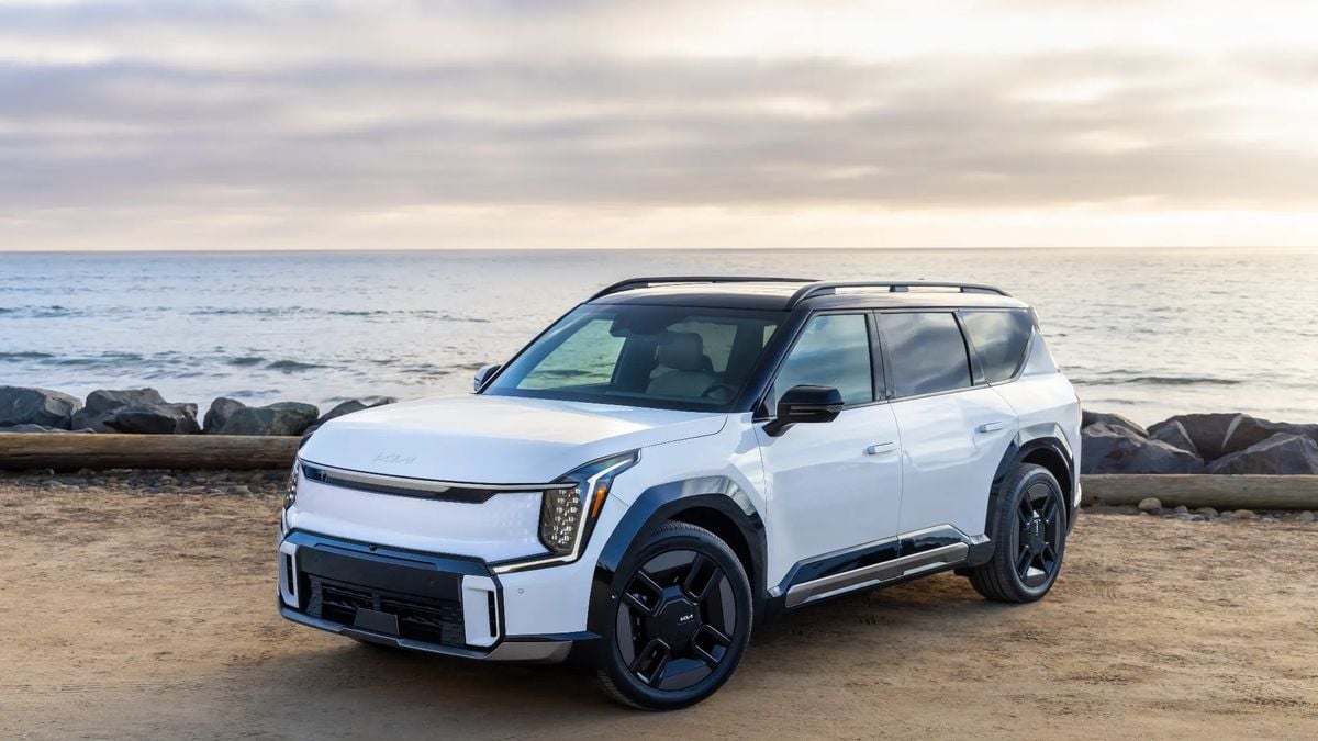 White Kia EV9 electric SUV parked on beach with ocean and rocky shoreline backdrop