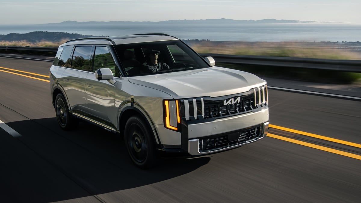 Cream-colored Kia Telluride HEV SXP driving on a coastal highway, front three-quarter view with ocean and hills in the background
