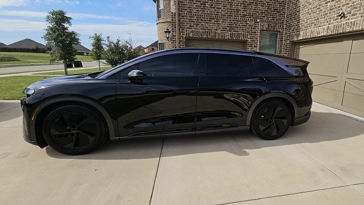 Side profile of a black 2026 Lucid Gravity GT parked outside a residential home with dark aero wheels and tinted windows.