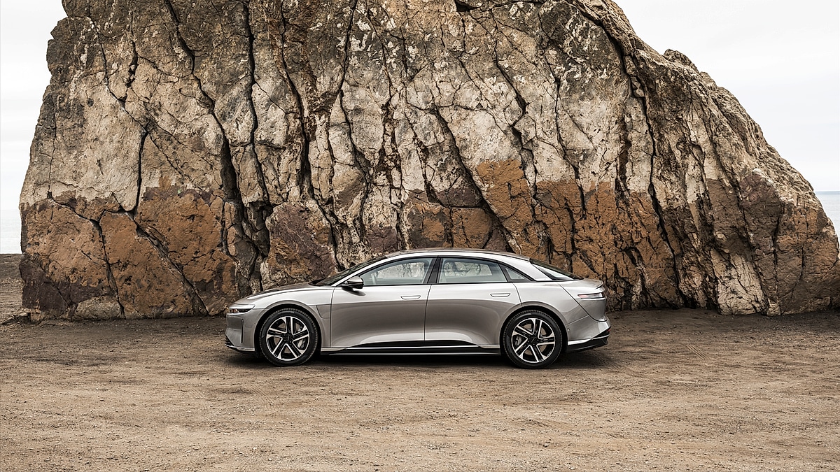 Silver Lucid Air shown in side profile beside a large coastal rock formation.