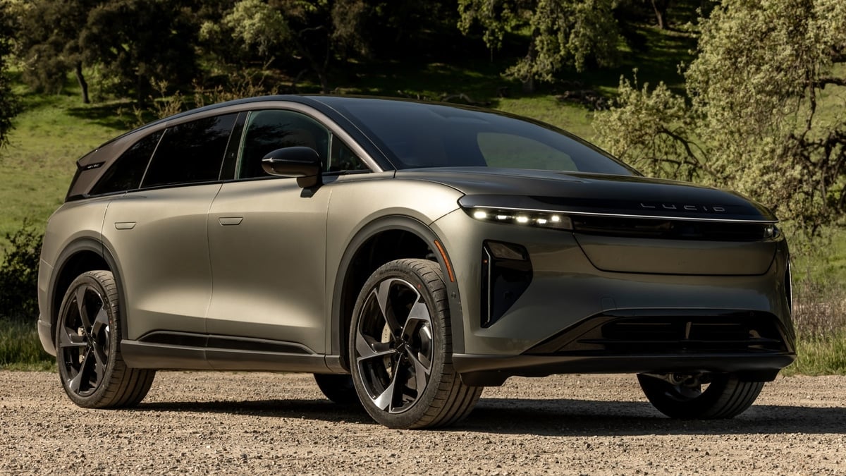 Silver Lucid Gravity electric SUV parked on a gravel road surrounded by oak trees, front three-quarter view showing headlights and grille
