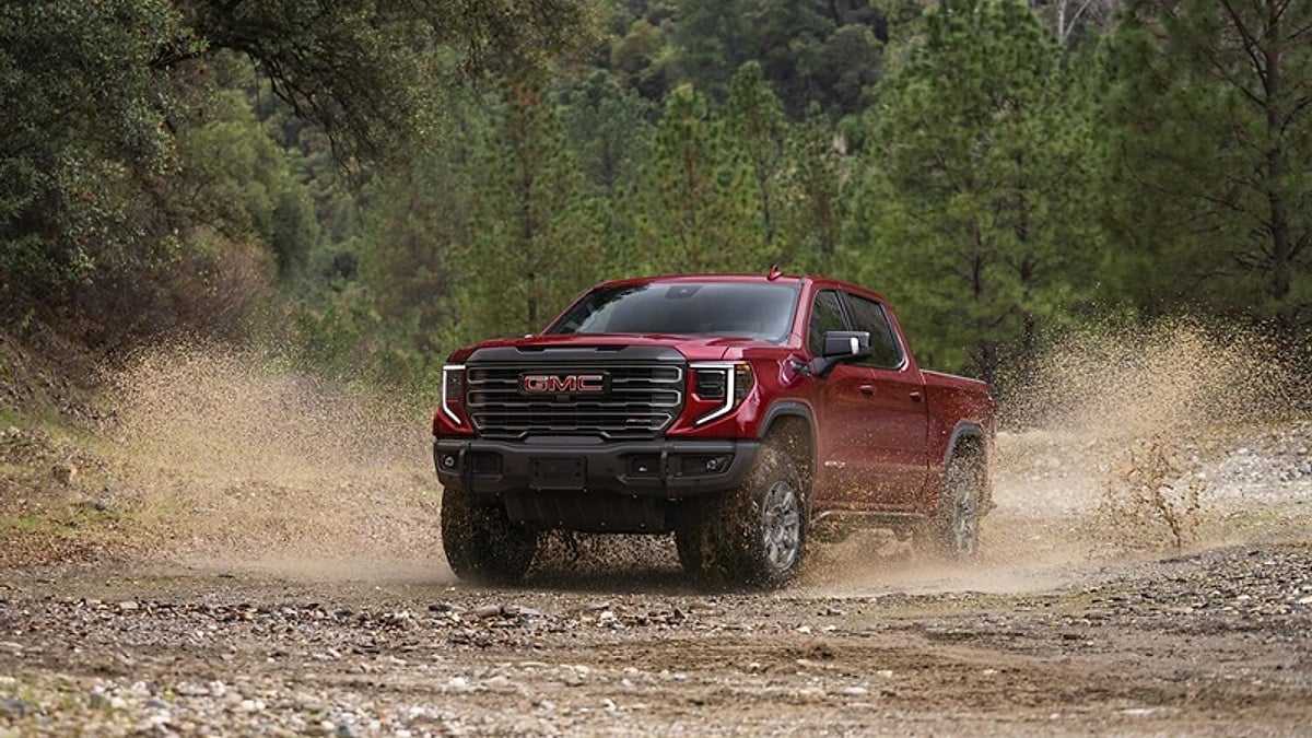 Red GMC Sierra 1500 pickup truck driving through mud and water on a forest trail.