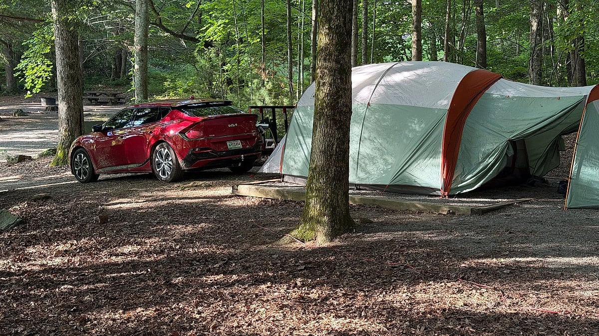 Red Kia EV6 parked beside a large tent at a wooded campsite.