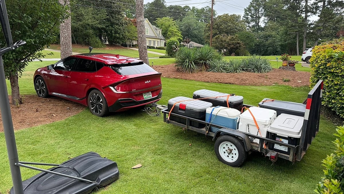 Red Kia EV6 towing a small utility trailer loaded with storage bins in a residential yard.