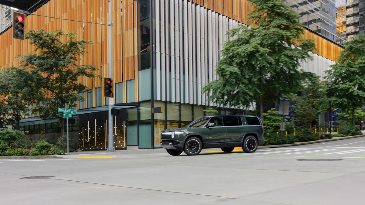 Dark green Rivian R1S SUV driving through a city intersection past a modern building with orange and white paneling