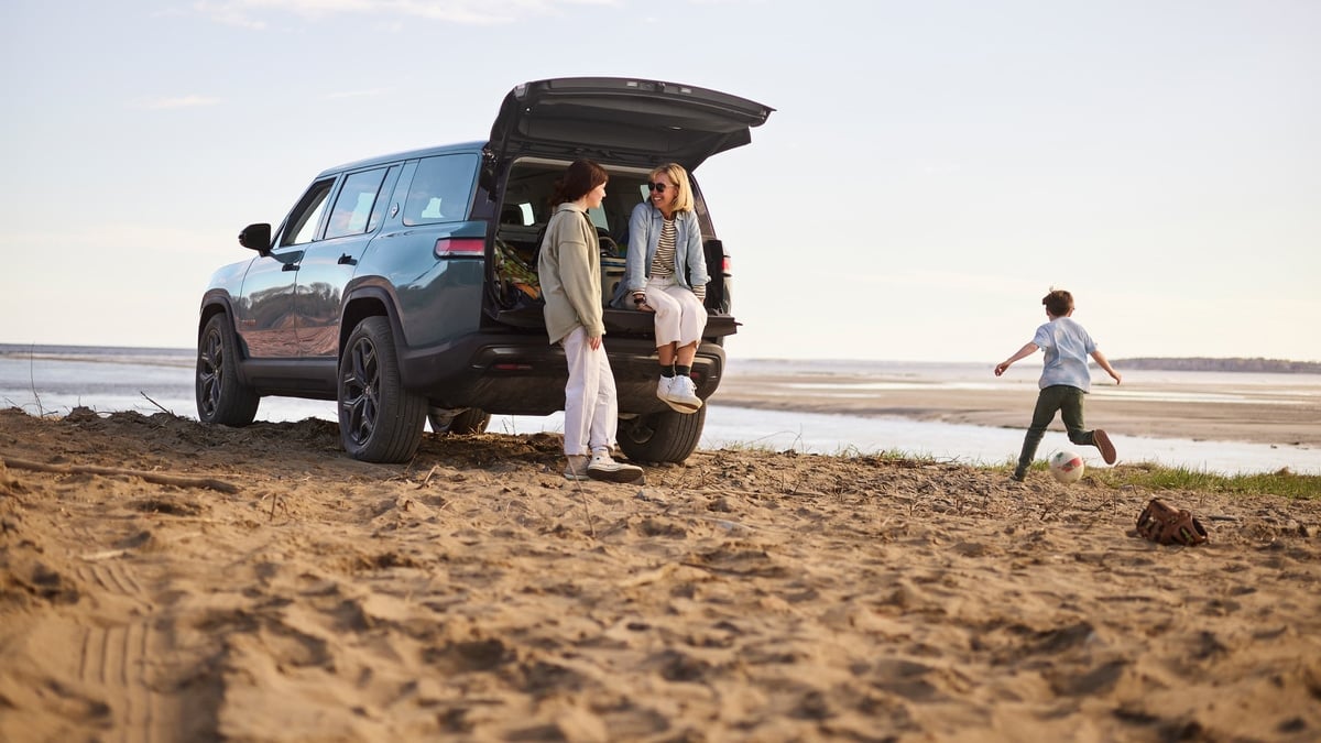 Family relaxing by the open trunk of a Rivian R1S electric SUV at a sandy beach during sunset.