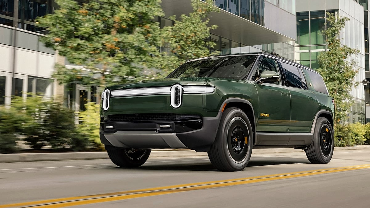 Green Rivian R1S driving past modern buildings in a front three-quarter urban action shot.