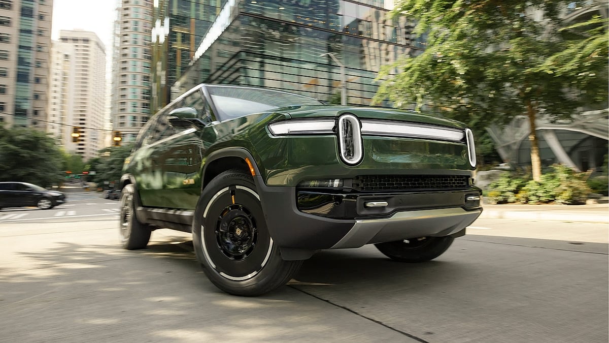 Green Rivian R1S electric SUV driving through a city street past glass buildings.