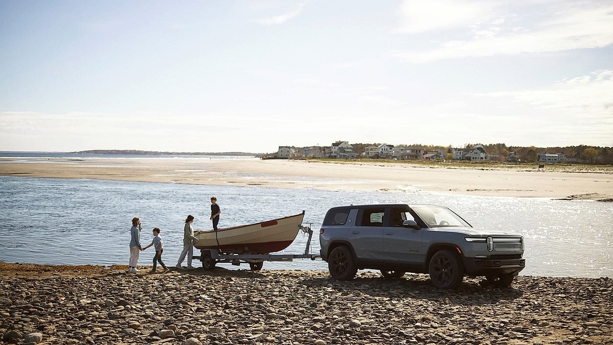 Rivian R1S parked on a rocky beach while towing a small boat, with a family standing nearby.