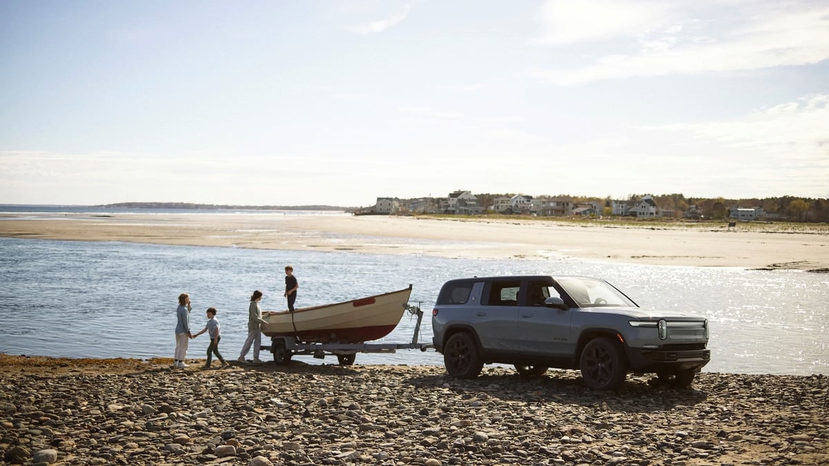 Gray Rivian R1S SUV towing a small wooden boat on a rocky beach, with a family unloading it near the water