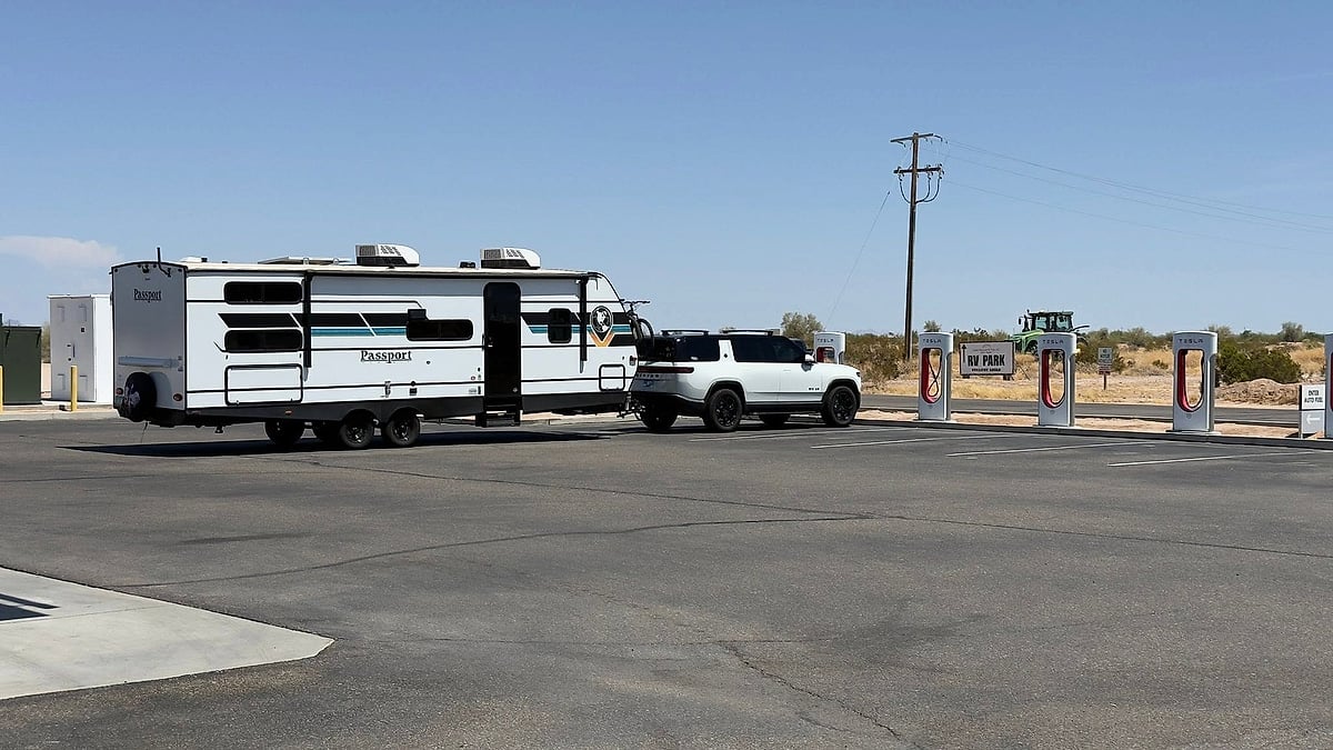 White Rivian R1S towing a Passport travel trailer near Tesla Supercharger stalls in a desert parking lot.